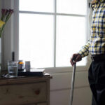 Healthspan Vs. Peakspan: The Battle for Vitality Elderly man standing with a cane next to a table with flowers and medication
