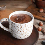 A cup of hot chocolate topped with chocolate shavings and surrounded by hazelnuts