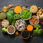 An assortment of legumes, nuts, and fresh vegetables arranged in bowls on a dark surface