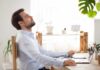 Man relaxing at desk with eyes closed beside laptop