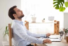 Man relaxing at desk with eyes closed beside laptop
