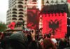 Crowd at an outdoor event with large red banners and a black double-headed eagle