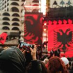 Crowd at an outdoor event with large red banners and a black double-headed eagle