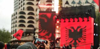 Crowd at an outdoor event with large red banners and a black double-headed eagle
