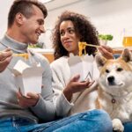 couple eating takeout noodles on a couch with a dog