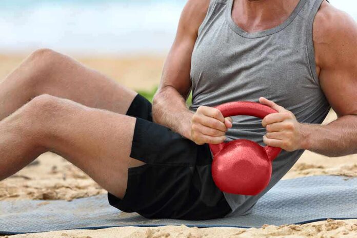 Man doing sit-ups with a red kettlebell on a beach mat